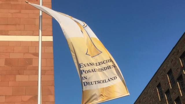 Eine Flagge mit dem Logo und Schriftzug „Evangelischer Posaunendienst in Deutschland“ weht an einem Mast vor blauem Himmel. Links ist eine Fassade aus rötlichen Steinblöcken zu sehen, rechts ein dunkleres Gebäude. Die Szene wirkt klar und sonnig.