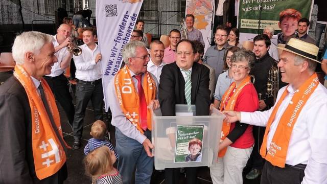 Eine größere Gruppe von Menschen steht bei einer öffentlichen Veranstaltung zusammen, einige tragen orangefarbene Schals. In der Mitte halten mehrere Personen gemeinsam eine transparente Box, während andere lächeln und zuschauen. Im Hintergrund sind Banner, Blasinstrumente und weitere Teilnehmende zu sehen.