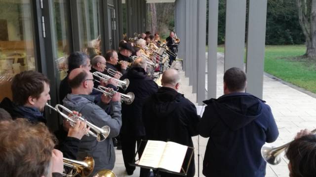 Ein Bläserensemble spielt unter einem überdachten Gang neben einem modernen Gebäude. Die Musiker stehen dicht nebeneinander mit Trompeten und Posaunen, vor ihnen liegt ein aufgeschlagenes Notenbuch. Im Hintergrund ist eine grüne Parkanlage zu sehen.