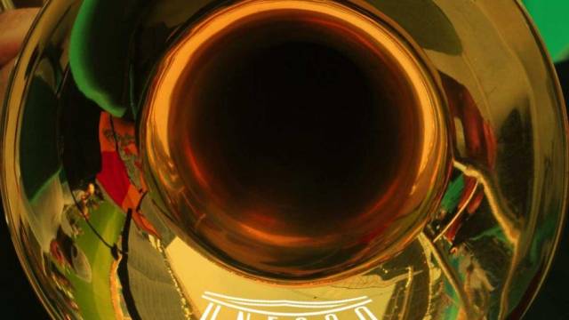 Nahaufnahme eines goldfarbenen Blechblasinstruments, der die Umgebung spiegelnd verzerrt. In der glänzenden Oberfläche sind Farben, Menschen und Gebäude reflektiert zu erkennen. Das Bild wirkt abstrakt und kunstvoll.