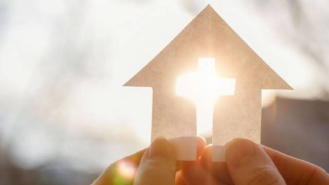 Ein gleichmäßiges Nahbild, auf dem zwei Hände ein ausgeschnittenes Papierhaus-Symbol vor eine Sonne halten — durch das Haus-Dach scheint das Sonnenlicht und wirkt wie ein Kreuz.