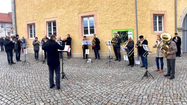 Ein Bläserensemble musiziert im Freien auf einem gepflasterten Platz vor einem gelben Gebäude. Die Musiker stehen im Halbkreis mit Notenständern, während ein Dirigent in der Mitte den Einsatz gibt. Alle tragen wetterfeste Kleidung, die Szene wirkt ruhig und konzentriert.