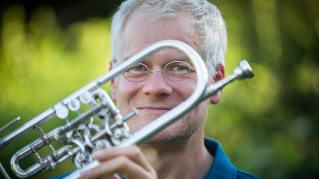 Nahporträt eines lächelnden Mannes mit kurzem grauem Haar und runder Brille; er hält ein silbernes Blechblasinstrument dicht vor das Gesicht, Hintergrund: grünes Laub, weich unscharf