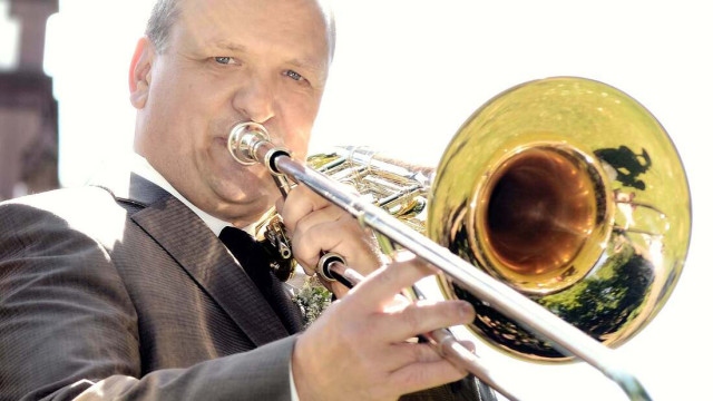 Nahaufnahme eines älteren Mannes im grauen Anzug und schwarzer Krawatte; er spielt eine Posaune, der goldene Schallbecher füllt die rechte Bildseite, Hintergrund sehr hell/überstrahlt