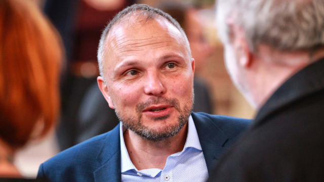 Porträt eines lächelnden Mannes mit Brille und grauem Haar im dunklen Sakko vor hellem, unscharfem Hintergrund