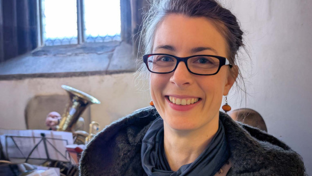 Porträt einer lächelnden Frau mit dunkler Brille und hochgestecktem, leicht zerzaustem Haar; sie trägt einen dunklen Schal und einen warmen Mantel, Hintergrund: Kircheninnenraum mit Spitzbogenfenster, Notenständern und Blechblasinstrumenten