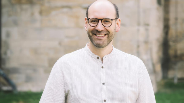 Nahporträt eines lächelnden Mannes mit Glatze, dunkler runder Brille und kurzem Bart; er trägt ein helles, fein gestreiftes Hemd mit Stehkragen, Hintergrund: helle Steinwand, weich unscharf