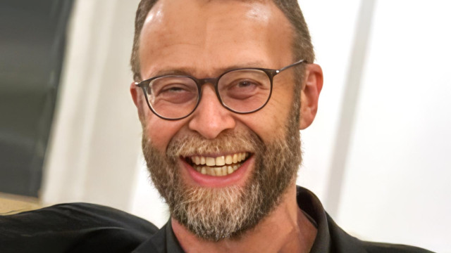 Nahporträt eines lachenden Mannes mit runder Brille und Vollbart vor hellem, unscharfem Hintergrund